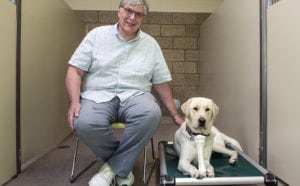 Bill sits in a chair in a suite in the canine center. He is smiling at the camera with one hand on the yellow lab lying next to him on a Kuranda bed