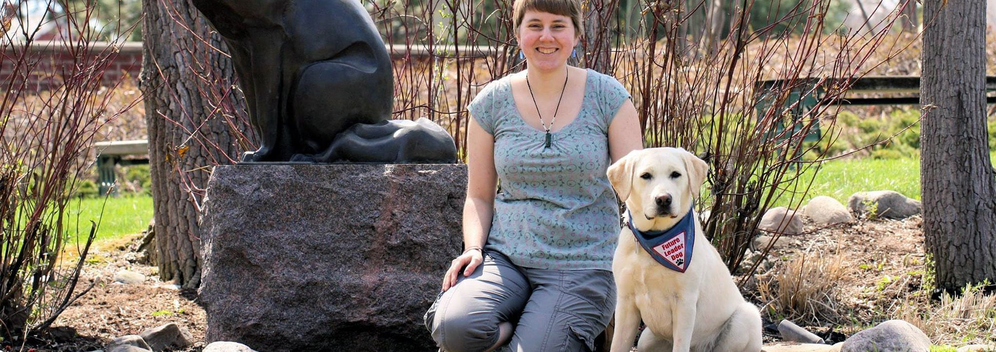 A young woman kneels on grass smiling at the camera with one hand on a seated yellow Labrador in blue Future Leader Dog bandanna next to her