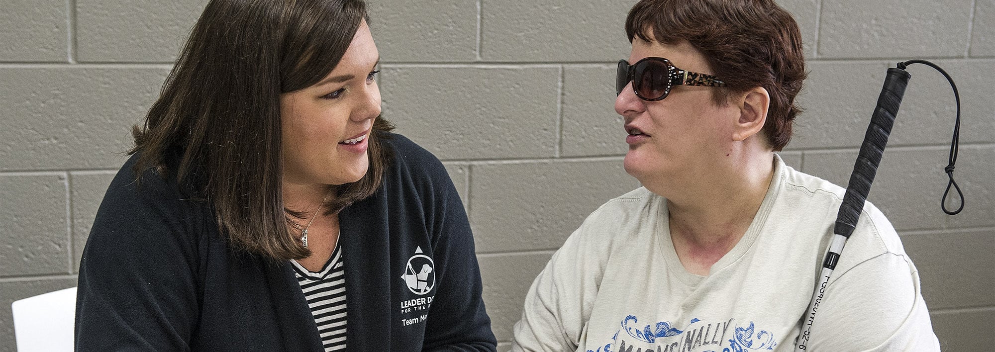 Two women sit talking face to face. One woman is holding a white cane