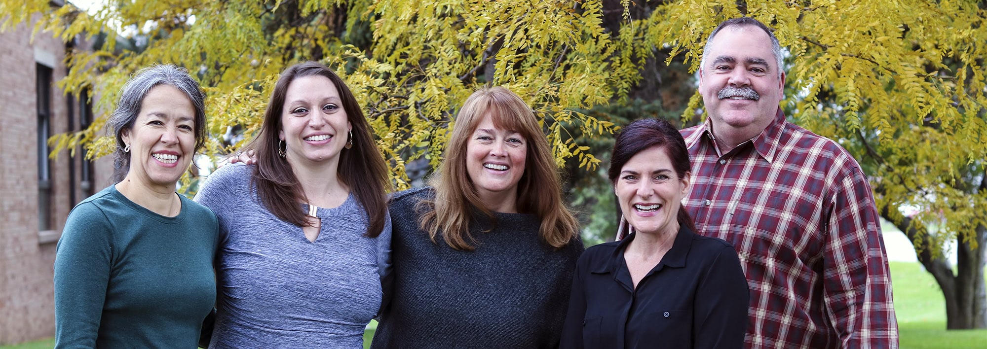 Ana, Melissa, Allison, Juliana and Jim of the client services team at Leader Dog stand with their arms around each other smiling at the camera in front of a tree