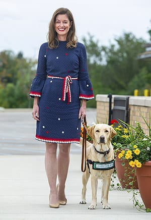 Melissa walks outdoors toward the camera, smiling, with a yellow Labrador ambassador dog in green ambassador jacket walking next to her on leash