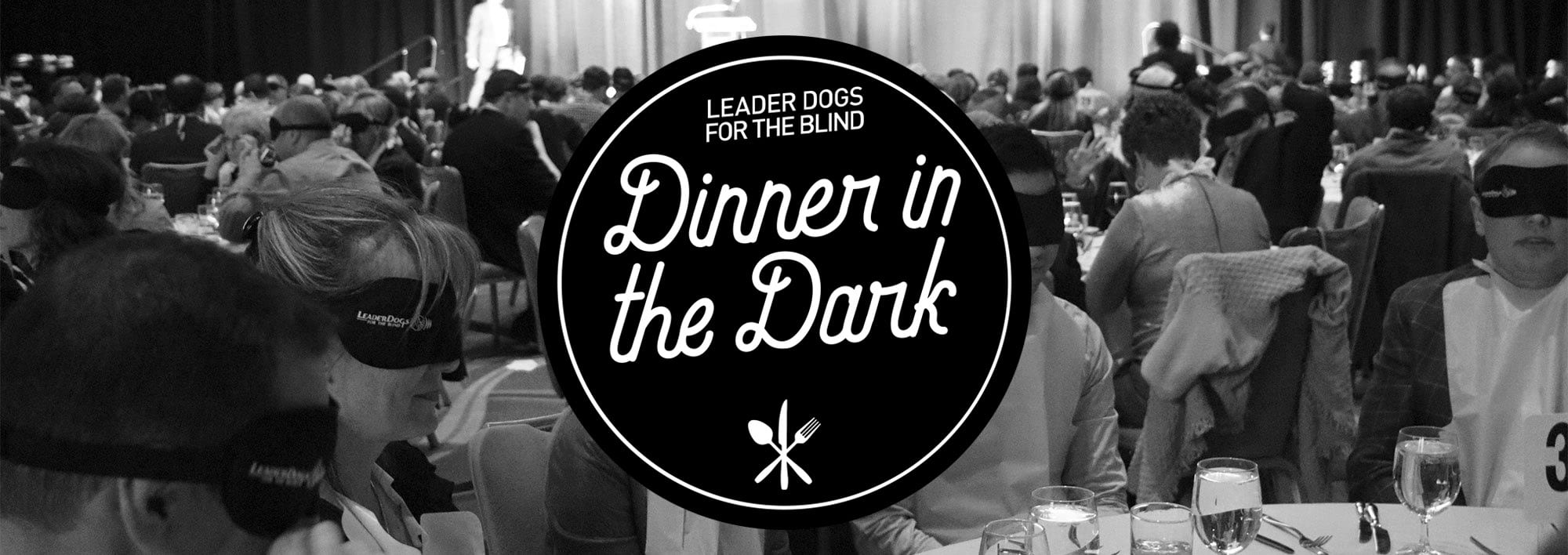 Black and white image of tables of people wearing blindfolds in a large room. There are drinks and food on the tables. In the center of the image is the black and white Dinner in the Dark logo