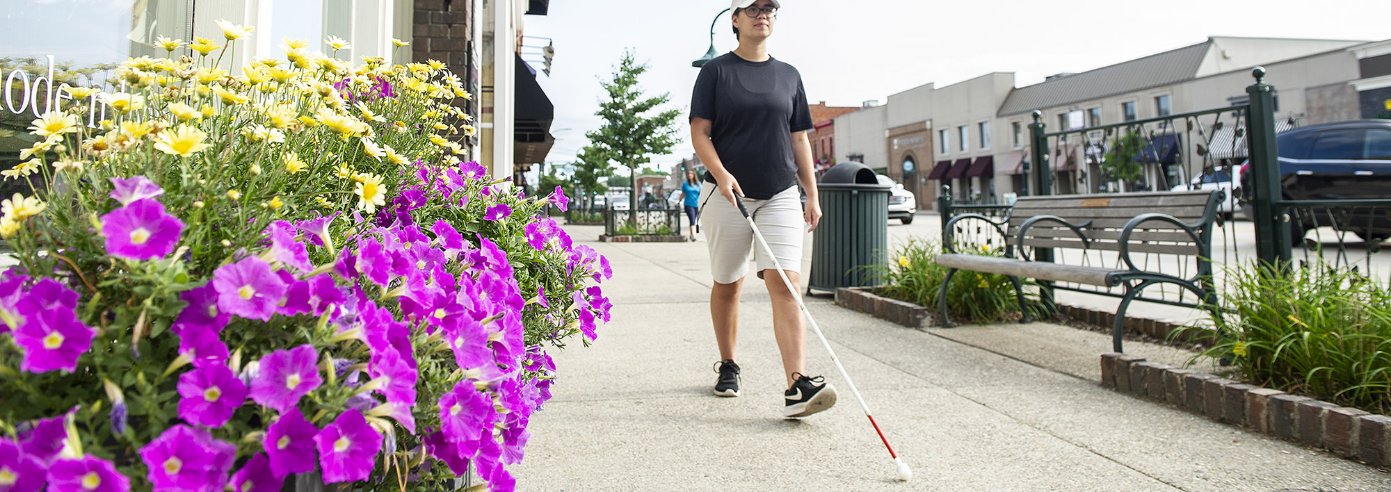 A young woman walks along a downtown sidewalk with traffic on her left side and flower boxes and a storefront on her right. She is using a white cane to navigate