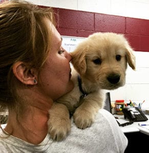 Ashley's back is to the camera. There is a golden retriever puppy peeking over her shoulder, looking at the camera. Ashley has her head turned toward the puppy.