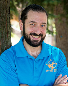 Tommy leans against a tree on a sunny day. He is smiling at the camera and wearing a bright blue polo with the Leader Dog logo on it in yellow.
