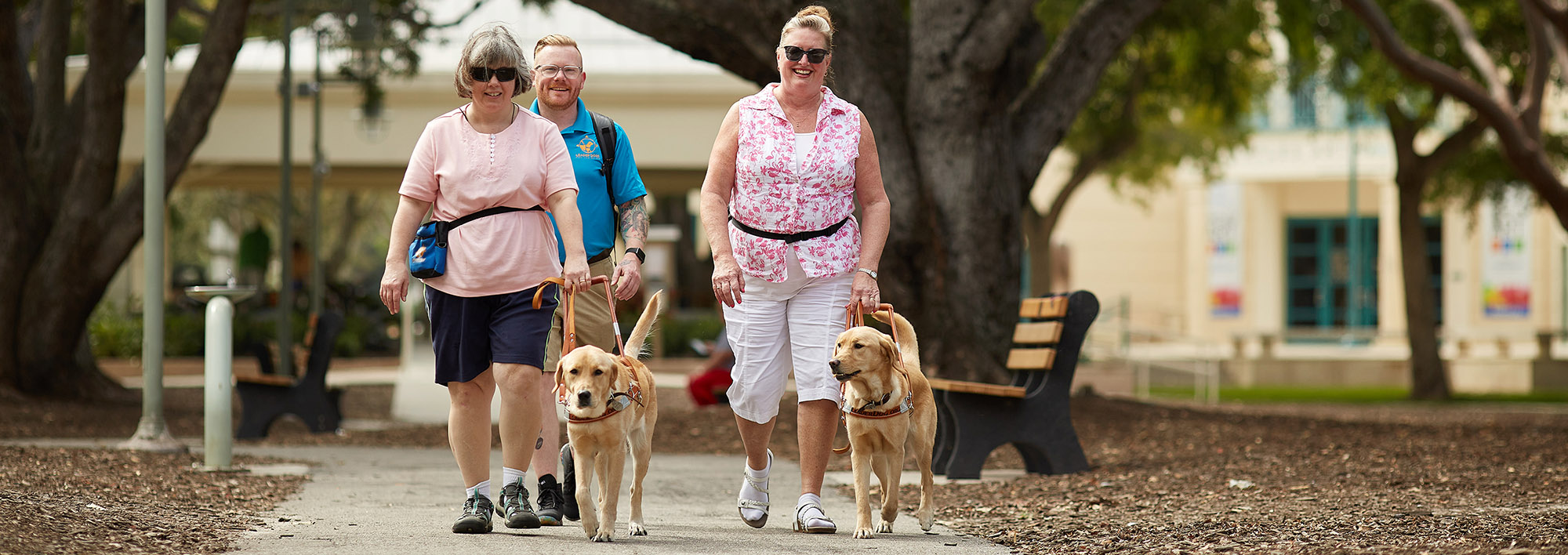 Two women walk down a sidewalk in a park-like setting toward the camera. They are smiling and both walking with Leader Dogs in harness. Behind them is a man wearing a bright blue polo with the Leader Dog logo on it