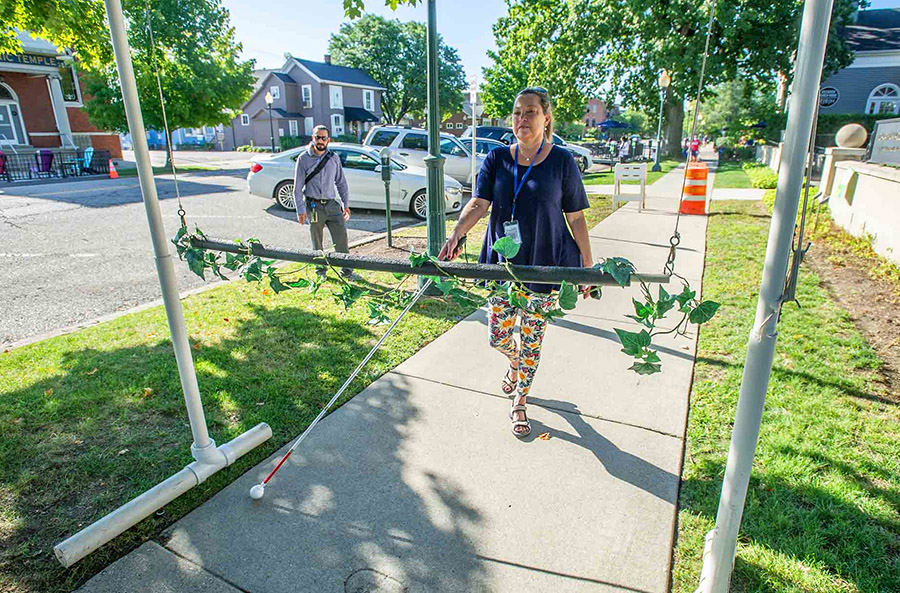 A woman with a white cane walks down a sidewalk on a sunny day. There is an arch with a bar through the middle placed on the sidewalk in front of her. This is an obstacle for her to practice working around.