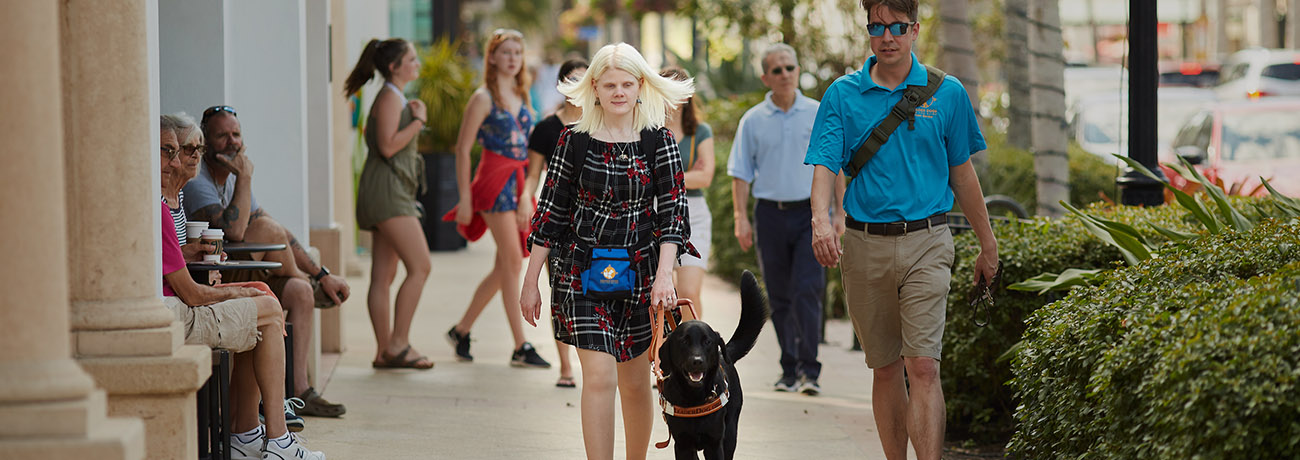 A woman in a black dress with a flowery print walks down a sidewalk toward the camera. She is holding the harness of a black lab Leader Dog. Her blonde hair streams out behind her. There are a number of people on the sidewalk around her, including a male Leader Dog instructor in a bright blue polo.