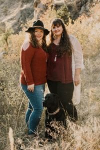 Cerr and her wife stand looking at the camera in a field of dry grass. Cerr is wearing an orange sweater, jeans, and a black hat. Katie is wearing a purple shirt and black pants with a white cardigan. They are both smiling. At their feet Iris the black Lab sits wearing a fall collar. She is looking off into the distance.