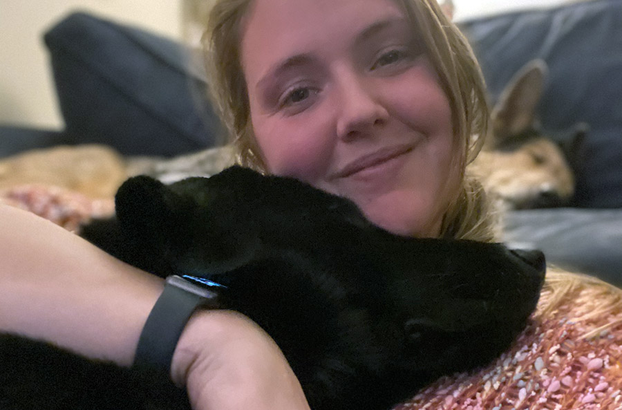 Ashley, smiling, hugs a black dog whose head is leaning against her.