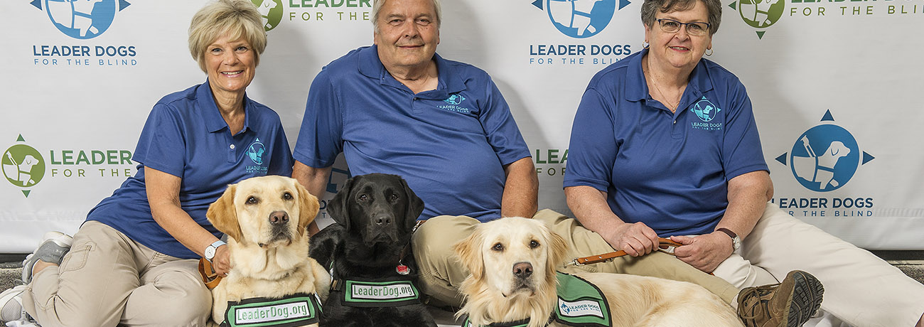 Two women and one man in blue Leader Dog polos sit on the floor with two labs (one black one yellow) and a golden retriever, all wearing green vests that say Leaderdog.org on the front strap