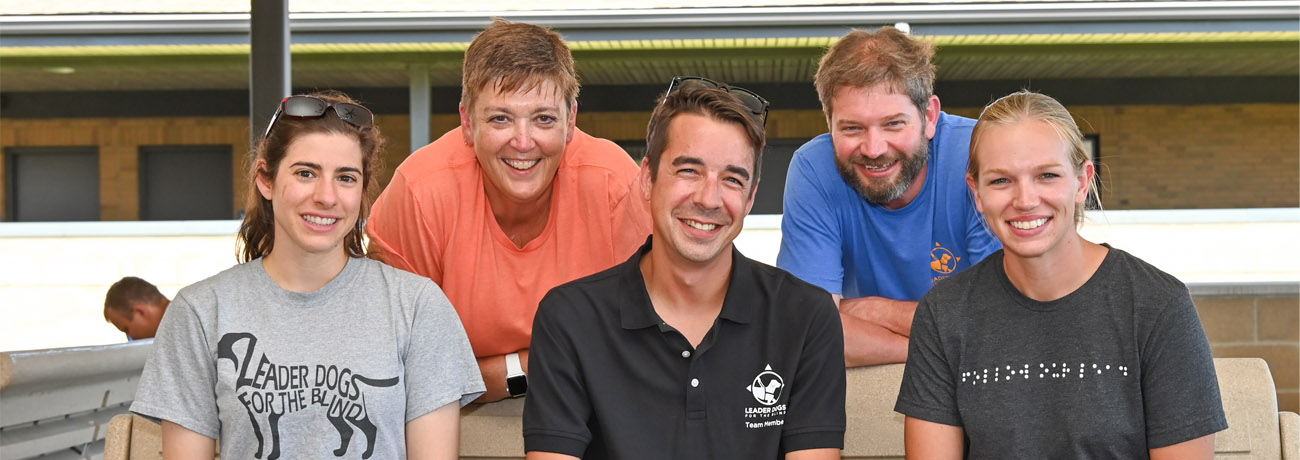 A team of Leader Dog instructors, three female and two male, smile at the camera. All are wearing assorted Leader Dog gear. Three are sitting on a bench with two leaning over behind it.