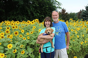 Karen and her husband with a young puppy in Karen's arms. They are smiling and standing in front of a field of sunflowers.