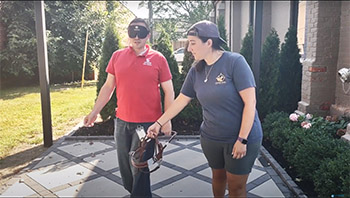 A man with a blindfold on holds a Leader Dog harness in his hand. The harness is also being held by a woman who is not blindfolded. They are outdoors.