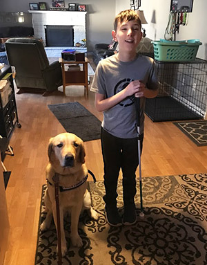 An older yellow lab/golden retriever puppy sits next to a young boy holding a white cane. They are in a living room.