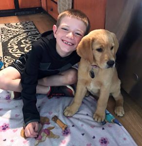 A young yellow lab/golden retriever puppy sits on a kitchen floor next to a young smiling boy with his arm around the puppy.