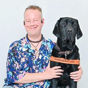 Jackie sits in front of a light grey photo backdrop, smiling, next to black lab Nico in his Leader Dog harness