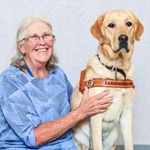 Sandy S sits smiling with yellow lab Mira in harness seated next to her