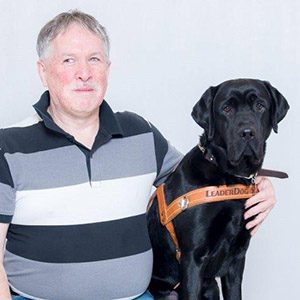 Tim looks toward the camera with his arm around black lab Leader Dog Ulric, seated next to Tim in harness