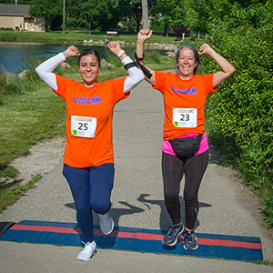 Two women cross a blue and orange line across a sidewalk with their hands in the air, smiling. They are wearing orange Bark & Brew 5K race shirts and bibs.