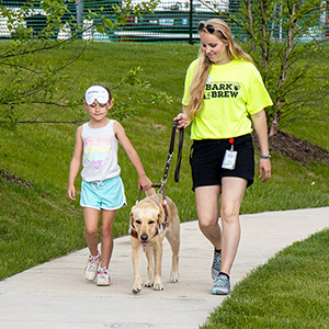 A blonde woman walks along a sidewalk holding the leash of a yellow lab. A young girl is walking next to the dog holding its harness. She has a blindfold pulled up over her eyes.