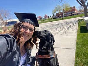 A selfie photo of the head and shoulders of a woman and a dog on a college campus. In the background are several large buildings and a large crowd of people. The woman has brown curly hair and glasses. She is wearing a black graduation hat and gown. She is leaning down to the black Labrador retriever who is wearing a light brown leather Leader Dog harness and dark brown leather collar. The dog seems to be sniffing the woman’s ear or hair.