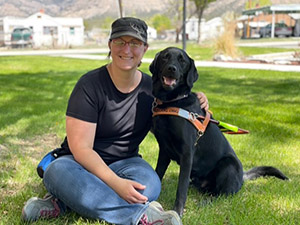 A woman and a black Labrador retriever sitting on the grass with some houses and mountains blurred for effect in the background. The woman is sitting cross-legged wearing jeans, tennis shoes, a black t-shirt, glasses and a black baseball cap. She is smiling and has her left arm around the dog. The dog is wearing a brown leather harness with LeaderDog.Org stamped on the front. He is sitting and looking forward with his mouth slightly open showing his pink tongue and a few white teeth. His eyes and nose are very black.