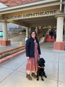 A smiling woman and a black Labrador retriever standing outside an event venue with the words “Tuacahn Amphitheatre” above the entrance. The woman has long slightly curly brown hair and is wearing glasses, brown boots, a red dress and a black coat. The dog is sitting at her left side and is wearing a light brown leather Leader Dog harness.