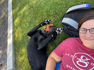 A woman and a black Labrador retriever are lying on their backs on the grass. The woman is wearing glasses and a red Garth Brooks concert t-shirt. Her head is resting on a black backpack and she is smiling. The dog is lying next to her with a chew toy in his mouth and his front legs in the air showing the pads of his feet.