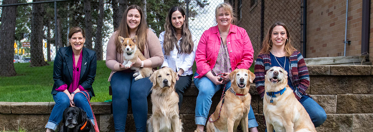 Five women, (Juliana, Danielle, Andy, Allison and Melissa) sit outside on a low brick wall with their dogs (black lab, corgi, golden retriever and 2 yellow labs)