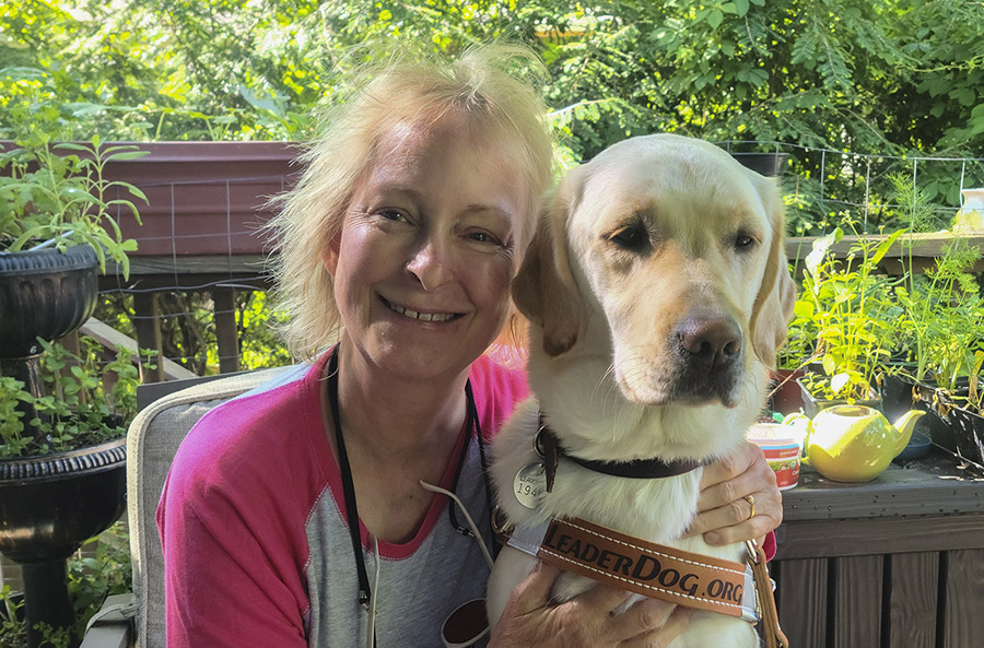 A smiling woman with short blond hair holds a yellow lab in Leader Dog harness next to her. Behind them is greenery