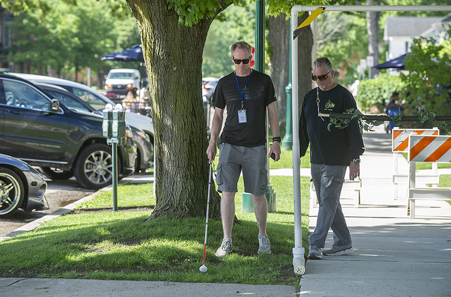 A man with a white cane walks on sidewalk toward camera. It's sunny and there's grass along the path. He is followed by another man.