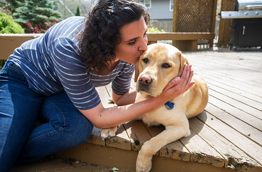 A dark-haired woman leans over on a sunny porch, hugging and kissing a yellow lab lying next to her