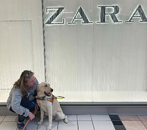 A woman and a dog are in a mall. The woman is squatting next to the dog, a yellow golden retriever/Labrador retriever cross, that is sitting on the tile floor. The woman is wearing jeans and a long sleeve shirt, she has long hair which is pushed back from her face with a pair of glasses used like a headband. The dog is wearing a brown leather Leader Dog harness and is looking off to one side. Behind the pair is a textured glass wall with the word “Zara” in all caps and lighted behind the glass.