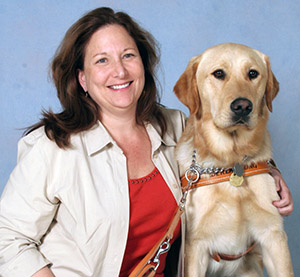 The first photo is of Helaine and her first Leader Dog, Chapman. They are sitting on a bench and Helaine has her left arm around Chapman’s shoulders and is holding his brown leather leash in her right hand. Helaine is wearing black slacks, a red shirt and a tan blouse, she has a large smile on her face. Chapman is a yellow Labrador retriever/golden retriever cross. He has dark eyes and a dark nose and is wearing a brown leather Leader Dog harness.