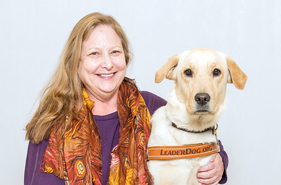 Helaine and her current Leader Dog, Zara. They are sitting on a bench and Helaine has her left arm around Zara’s shoulders. Helaine is wearing black slacks, a purple long-sleeve shirt and a brightly patterned yellow, tan and red scarf; she has a large smile on her face. Zara is a yellow golden retriever/Labrador retriever cross. She has dark eyes and a dark nose. She is leaning into Helaine and is wearing a brown leather Leader Dog harness.